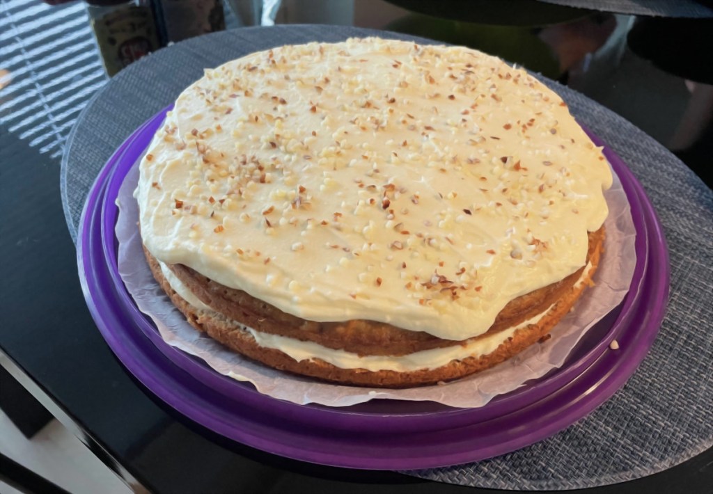 A large carrot cake on a plate. The carrot cake has a cream-colored icing and there are crushed hazelnuts sprinkled on top.