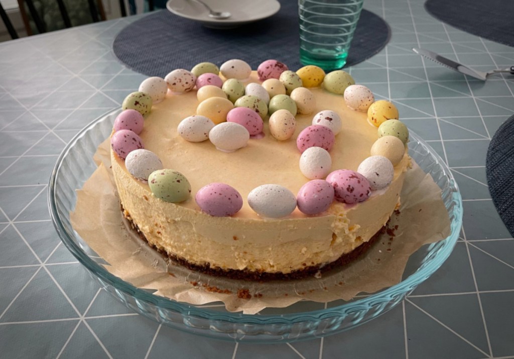 A round cheesecake on a plate is placed on a table that has plates and glasses set. The cheesecake has a thick, light yellow filling and it's decorated with small yellow, white, green and pink candies that look like miniature bird eggs.