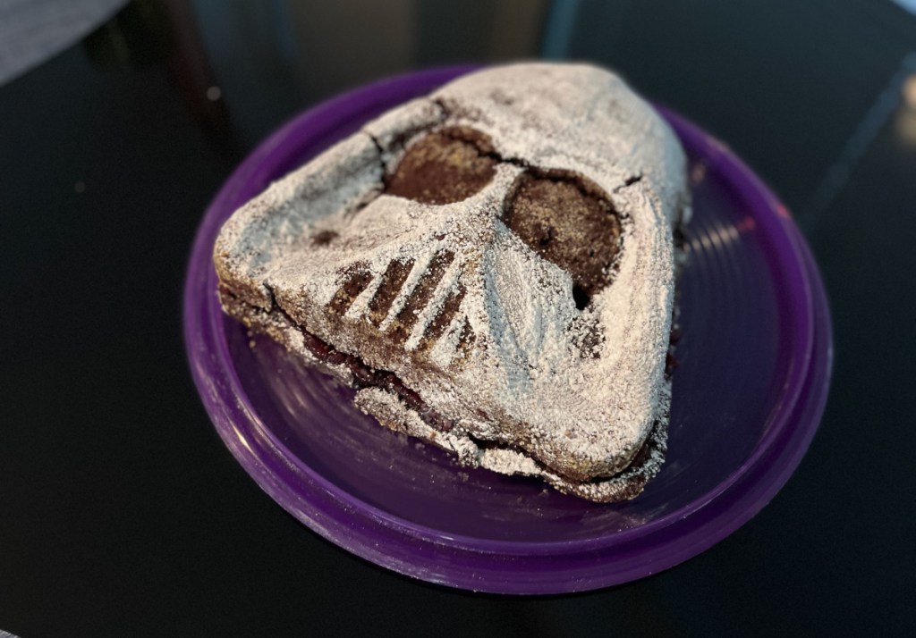 A chocolate cake shaped like Darth Vader's head is on a serving plate. The cake is covered with white powdered sugar. The eyes and the slits of the breathing mask are dark brown. 