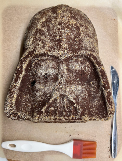 A brown undecorated cake shaped like Darth Vader's helmet on top of baking paper.